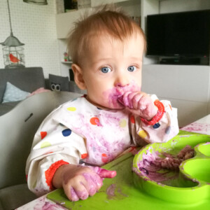 dziecko w ubrudzonym fartuszku siedzące w krzesełku do karmienia, ubrudzone jedzeniem, na stole stoi zielony talerzyk z przegródkami