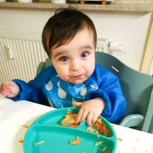 dziecko siedzące w krzesełku do karmienia, na stole zielony talerzyk z przegródkami i jedzeniem